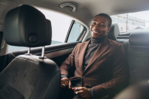 Young smiling african man using smartphone while sitting on backseat in car. Concept of happy business people traveling.